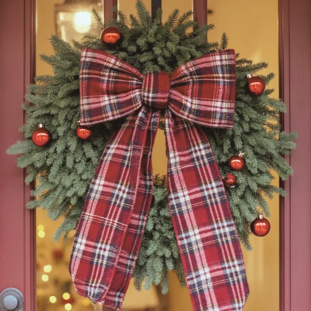 A flannel wreath with evergreen branches, red ornaments, and a large red plaid bow with long tails, on a glass front door.