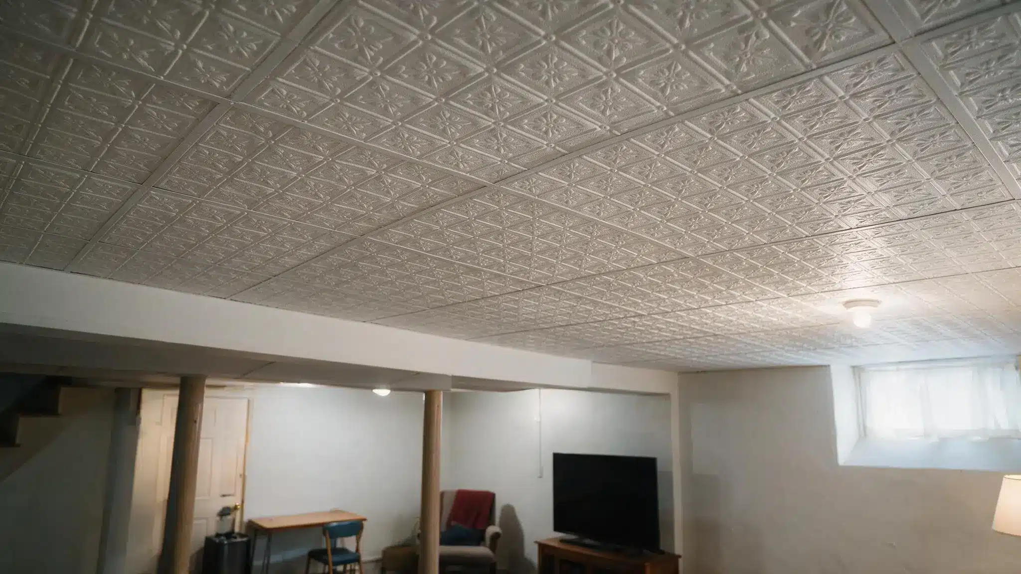 White patterned ceiling tiles above a basement room with a TV, table, chairs, armchair, and small window near the ceiling.