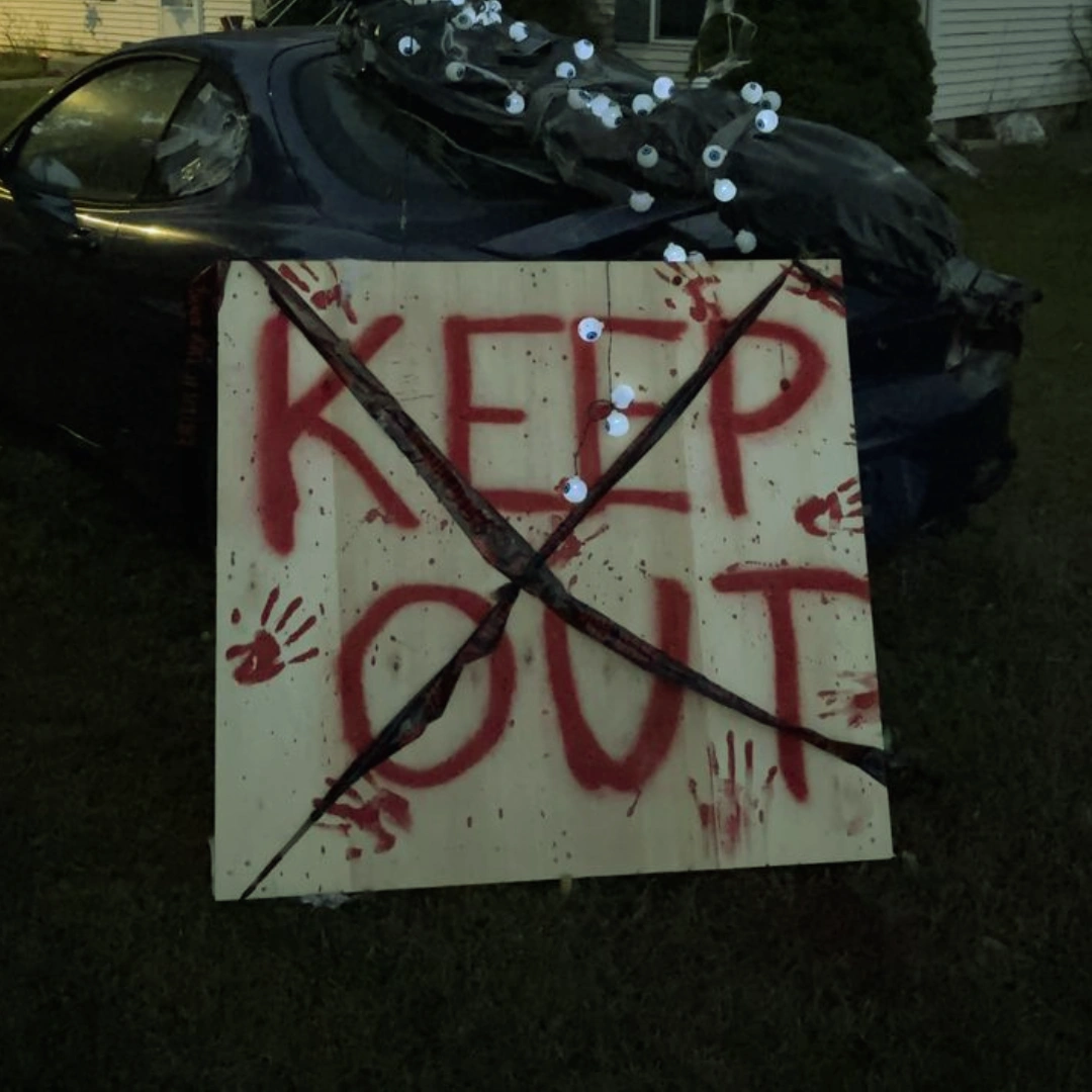 Halloween keep-out sign with red handprints and crossed branches, displayed in front of a dark car covered with eerie eyeball props.