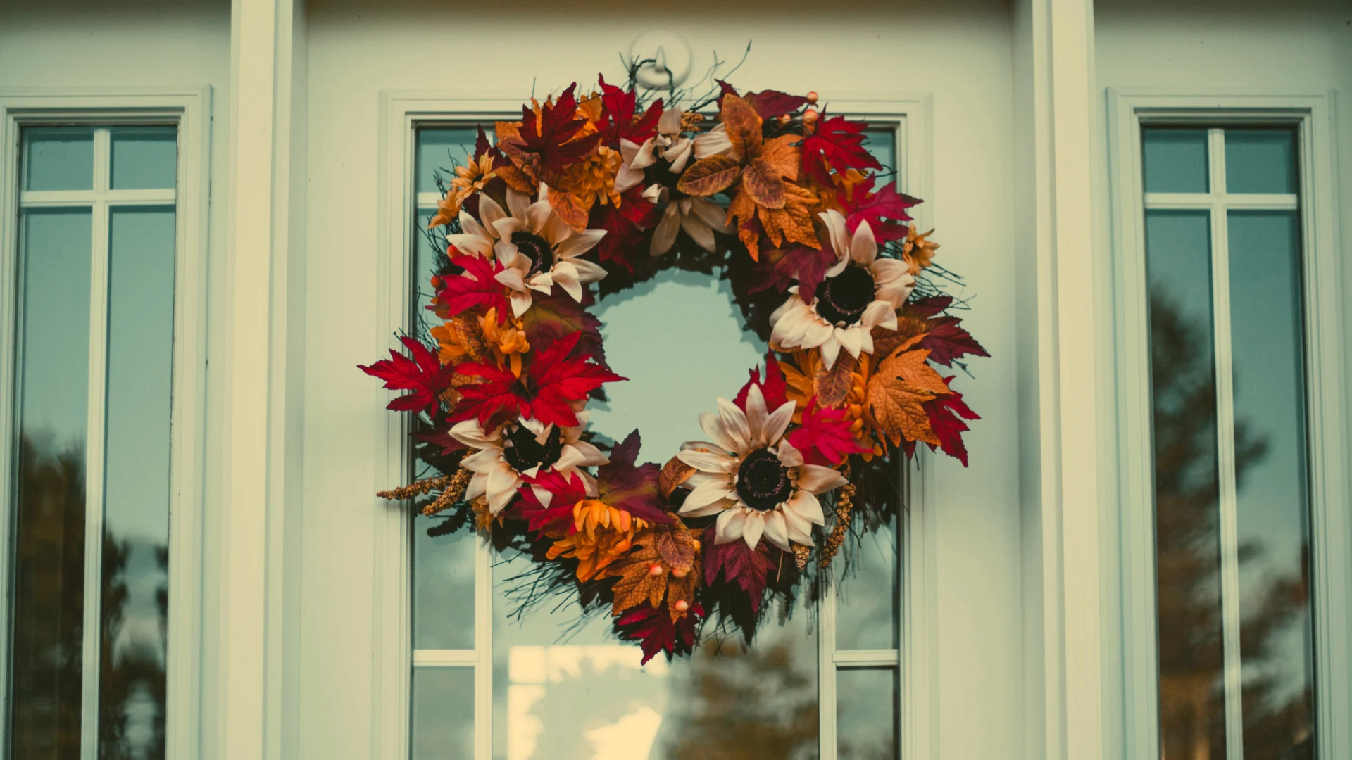 DIY fall wreath with red and orange leaves, cream sunflowers, and seasonal flowers hanging on a white glass-paneled front door.