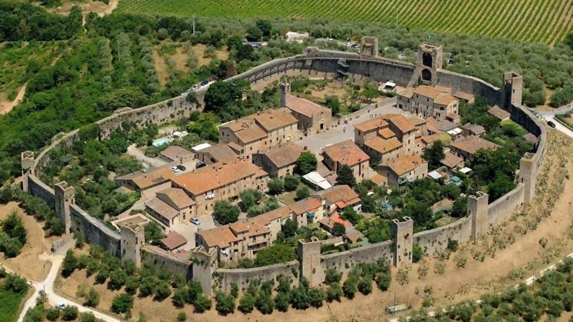 Aerial view of a medieval fortified town surrounded by stone walls and towers, with terracotta-roofed buildings and green vineyards.