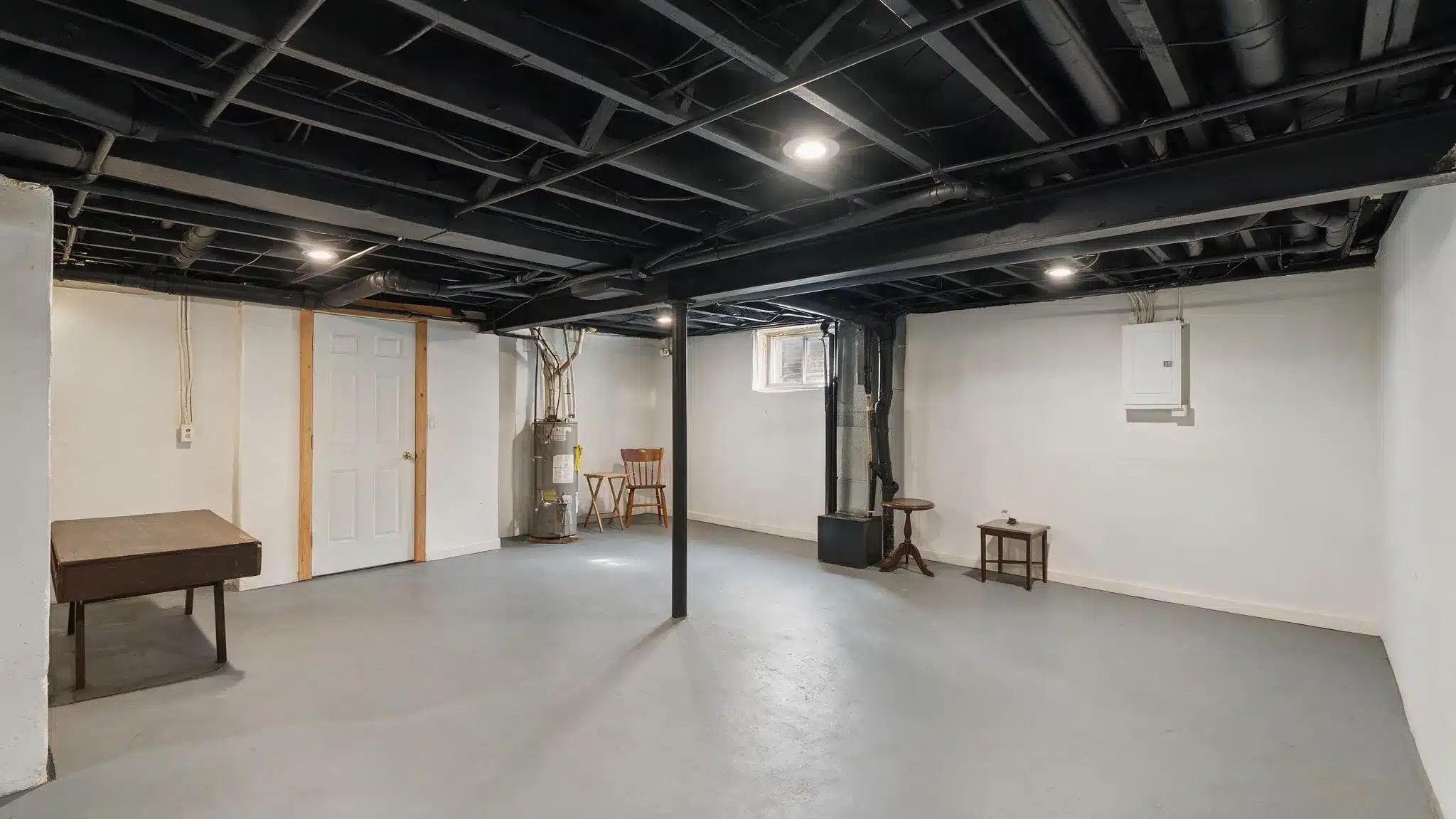 Unfinished basement with black-painted exposed ceiling, white walls, gray floor, a water heater, a door, and a few small tables.