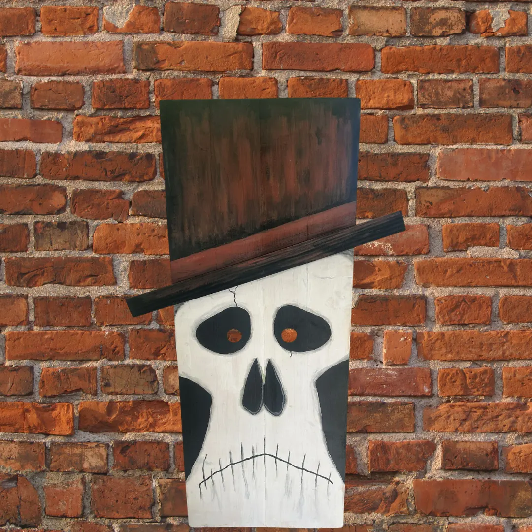 Wooden Halloween skull decoration with a tall black top hat and painted white face displayed against a rustic red brick wall.