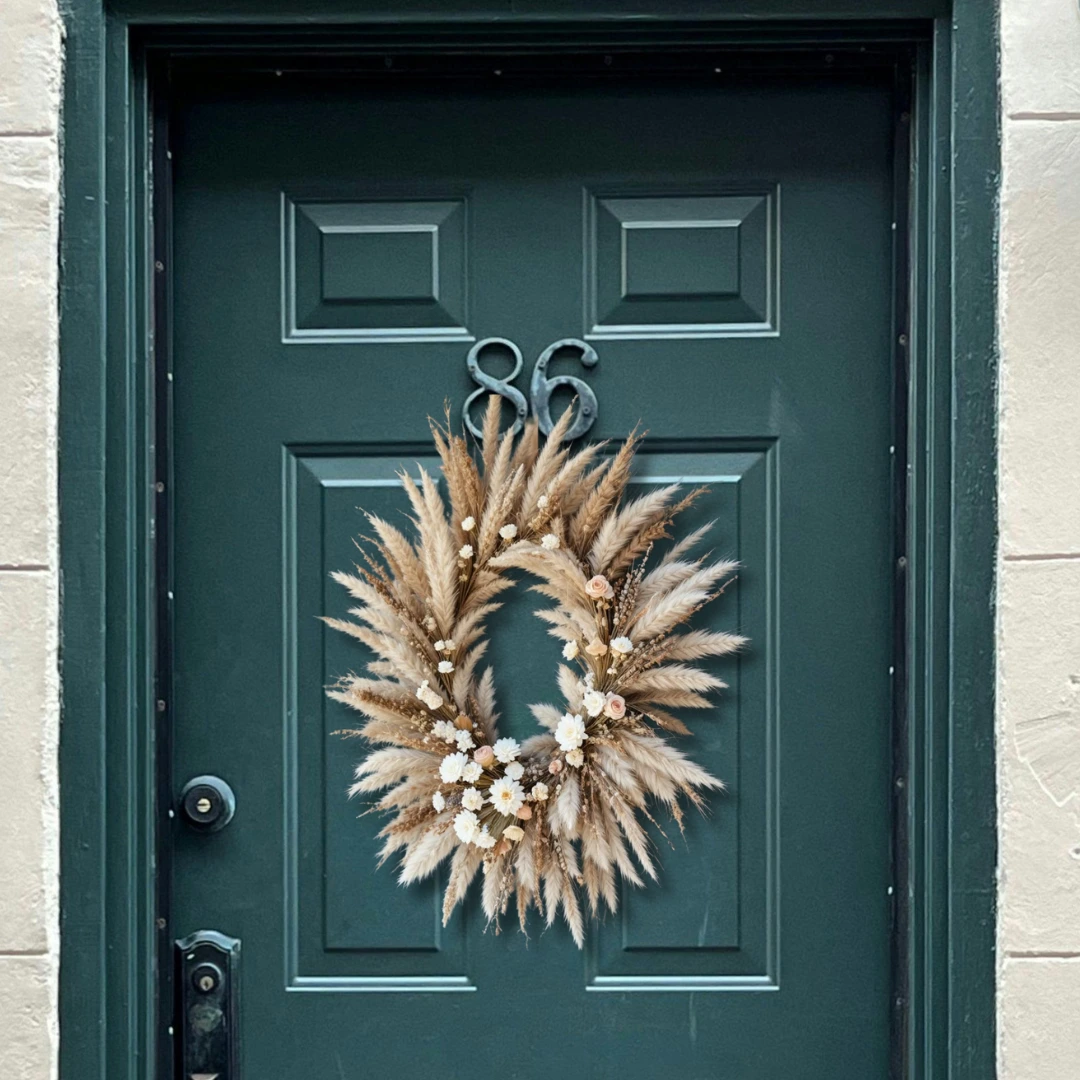 DIY fall wreath made with dried wheat, pampas grass, and small white flowers, hanging on a dark green paneled front door.