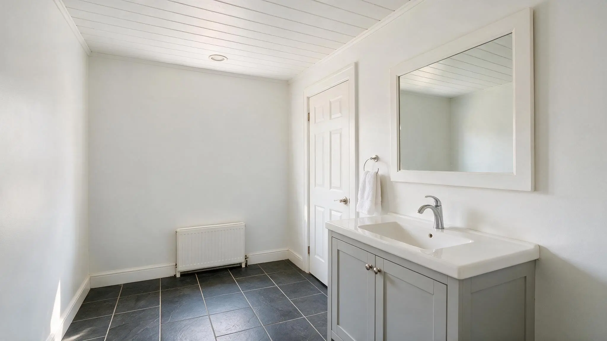 A clean white bathroom with a shiplap ceiling light walls a simple vanity large mirror and dark tile flooring