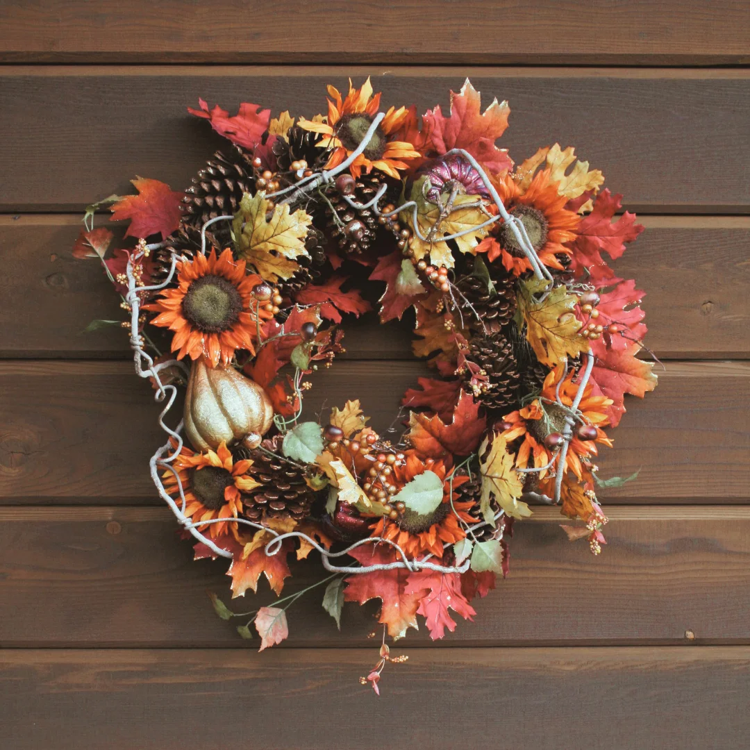 DIY fall wreath with orange sunflowers, red and yellow maple leaves, pine cones, berries, and gourds on a wooden wall.