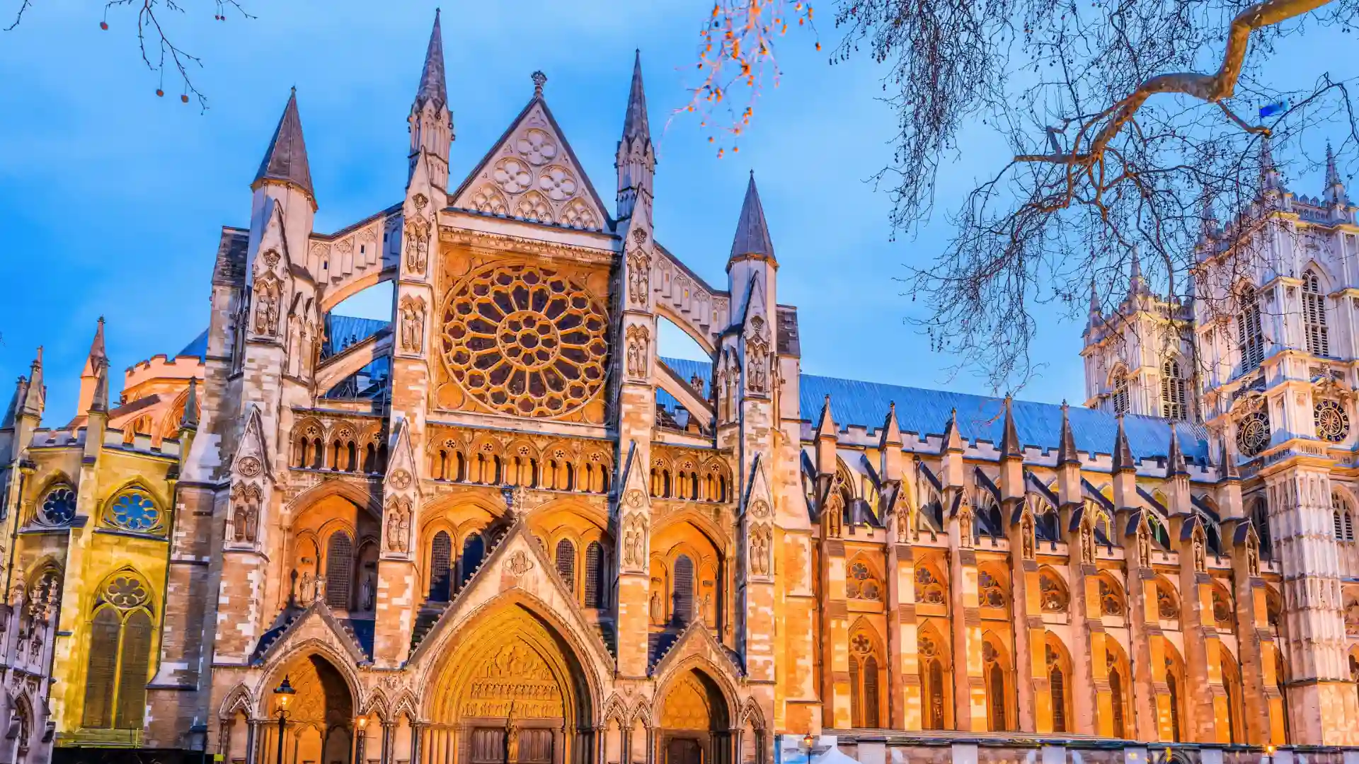 Westminster Abbey in London with ornate Gothic architecture, pointed spires, and a large circular rose window illuminated at dusk.